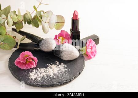 Pennelli cosmetici e polvere per il viso su un'ardesia, rossetto rosa rosso e teste di fiori su sfondo bianco, concetto di bellezza e trucco, spazio copia, selecte Foto Stock