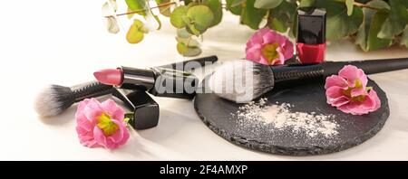 Utensili per il make-up come rossetto, smalto per unghie, spazzole e polvere per il viso con fiori rosa su un'ardesia nera e uno sfondo di legno bianco, bellezza e cosm Foto Stock