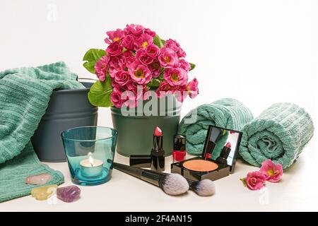 Disposizione degli utensili di bellezza e trucco come rossetto, smalto per unghie e polvere per il viso con spazzole cosmetiche, tovaglioli turchesi, candela e fiori rosa Foto Stock