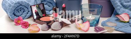 Utensili per il trucco come polvere per il viso, rossetto rosa rosso e smalto per unghie con spazzole cosmetiche, asciugamani blu, petali di candela e fiori, banner panoramico fo Foto Stock
