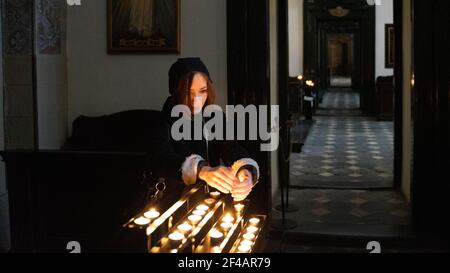 Una giovane donna caucasica che tiene le mani sopra il tè rotondo luce che brucia candele nella chiesa cattolica interior.in Cracovia, Polonia. Foto Stock