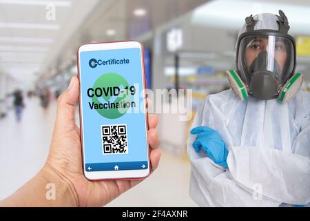 Smartphone con mano che mostra la vaccinazione Covid-19 sullo schermo con la donna ufficiale in tuta di protezione in aeroporto che si accaparra sullo sfondo. Foto Stock