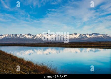 Neve coperta catena montuosa delle Alpi meridionali vicino a Twizel riflesso L'acqua ferma nel canale che esce dal lago Rautaniwha sotto un po' di nuvola spia Foto Stock