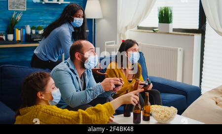 Amici tristi perché perdere di giocare ai videogiochi con controller wireless indossando la maschera facciale per evitare di ammalarsi con corona in tempo di pandemia sociale. Gruppo di persone che si divertono insieme. Foto Stock
