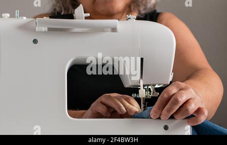 Primo piano frontale di una macchina da cucire, con una donna caucasica più anziana e irriconoscibile sullo sfondo, che sta facendo il lavoro di cucito. Foto Stock