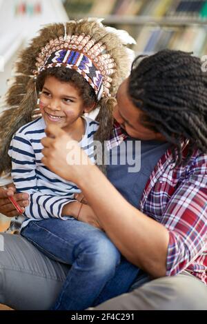 Un ragazzino vestito come un indiano seduto nel grembo del padre che ha fatto i colori indiani sul suo viso in un'atmosfera giocosa a casa. Famiglia, casa, playti Foto Stock