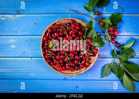 Ciliegia in ciotola di legno e rametti di ciliegie rosse mature su un tavolo di legno blu. Frutto di ciliegio biologico maturo, quadro piatto Foto Stock