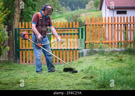 Uomo anziano falciatura erba da decespugliatore in giardino in primavera. Vecchio coltivatore tagliando erba. Foto Stock