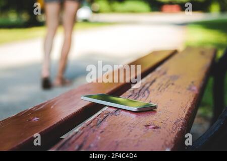 Smartphone smarrito sulla panchina nel parco pubblico. Donna che esce dalla panchina quando dimentica il suo smartphone. Foto Stock