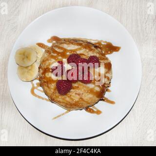 I gustosi pancake con sciroppo di caramello, lamponi e alcune fette di banana serviti su un piatto bianco Foto Stock