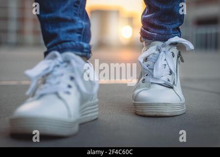 Sneaker bianche in città al tramonto. Camminare con le scarpe sportive sulla strada. Vista frontale ravvicinata delle calzature all'aperto. Foto Stock