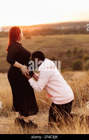 Felice coppia di matrimonio al campo, sposo sopravorato bacio incinta tummy di bella moglie, giovane famiglia si aspettano per un bambino, eccitato circa la paternità Foto Stock