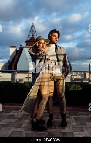 Coppia elegante avvolta in coperta stand al balcone al tramonto, bell'uomo abbraccio donna splendida, marito amorevole godere di tempo con bella moglie Foto Stock