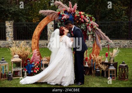 Bella donna bianca con affascinante afroamericano uomo stand vicino arco decorato, adorabile coppia in cerimonia di nozze, attraente bacio sposo bello Foto Stock