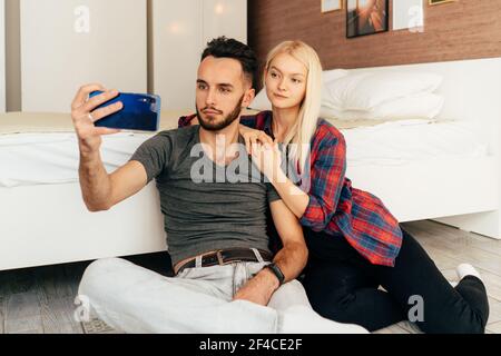 Una giovane coppia seduta sul pavimento nella stanza fa un selfie su un telefono mobile. Foto Stock