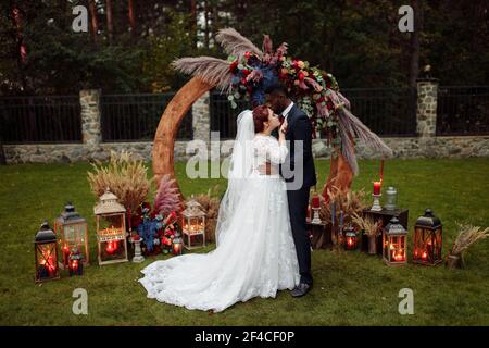 Bella coppia nella cerimonia di nozze, bell'uomo afroamericano con adorabile donna bianca stare vicino ad arco decorato, coccolarsi, godere felice Foto Stock