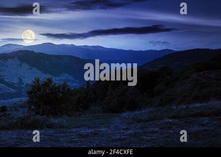 paesaggio montano rurale di notte. prato erboso in cima ad una collina. nuvole sopra la cresta in piena luce lunare. vista nella valle lontana Foto Stock