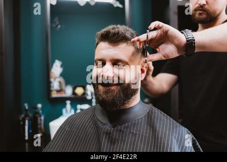 Felice cliente maschile soddisfatto di barbiere seduto in sedia mentre il parrucchiere gli dà i capelli Foto Stock
