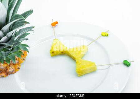 Spiedini di ananas sbucciati e tagliati a triangoli con bastoncini decorati su piastra bianca con foglie di ananas su sfondo bianco Foto Stock