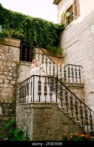 Una sposa con una corona di rose bianche e rosa si erge sulla scala con una ringhiera in legno battuto nella Città vecchia di Cattaro Foto Stock