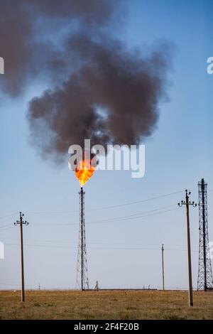 Torcia a gas naturale in fiamme di impianti di raffineria di petrolio in un deserto contro cielo blu e silhouette di tubo di fabbrica. Foto Stock