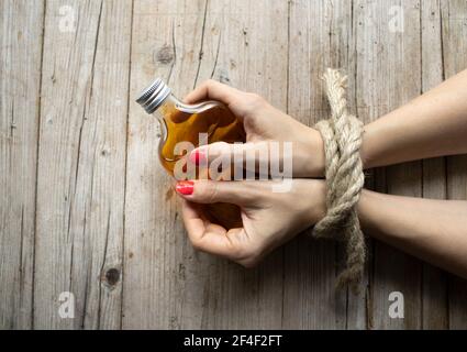 Mani di giovane donna con le unghie rosse sono legate e sdraiati su sfondo di legno chiaro, sta tenendo una piccola bottiglia di alcol, concetto di dipendenza Foto Stock