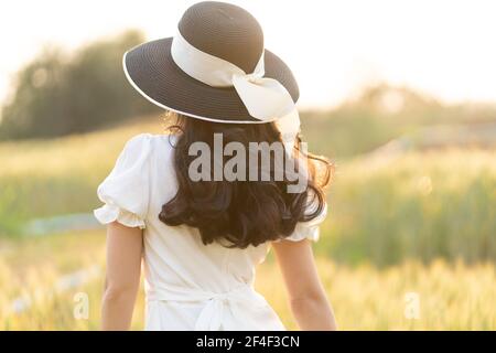 Vista posteriore di una giovane donna che indossa cappello nero e abito bianco camminando da sola in un campo d'orzo in un tardo pomeriggio prima del tramonto, calda luce d'ora d'oro a Foto Stock