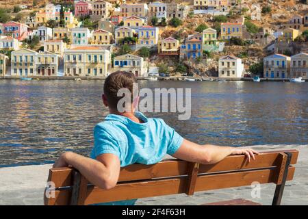 Giovane turista maschile seduto su una panchina e guardando il porto di Symi, Grecia. Concetto turistico. Visite turistiche. Destinazione estiva di vacanza. Viaggi conce Foto Stock