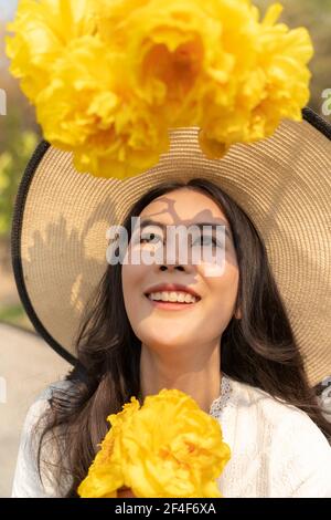 Ritratto di felice giovane bella donna con grande cappello di paglia e vestito bianco, sorridente e guardando in su grande fiore giallo davanti a lei su luminoso s. Foto Stock
