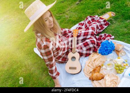 Ragazza in abito a scacchi rosso e cappello seduta su coperta da picnic in maglia bianca gioca ukulele e bere vino. Picnic estivo in giornata di sole con pane, frutta, fiori di bouquet idrangea. Messa a fuoco selettiva Foto Stock