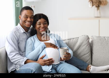 Felice coppia nera incinta coccolarsi sul divano a casa Foto Stock