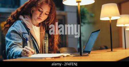 Ragazza che prende appunti nel suo libro sotto lampada in biblioteca, con computer portatile sulla scrivania. Giovane donna nella biblioteca universitaria che completa il suo incarico. Foto Stock