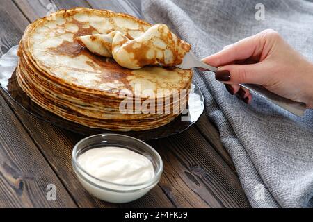 Una giovane donna raccoglie una frittella con una forchetta. Panna acida per pancake sul tavolo, colazione per tutta la famiglia. Maslenitsa, la vacanza della fine Foto Stock