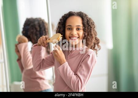 Allegra ragazza nera che combatte i suoi capelli ricci, preparandosi al mattino Foto Stock