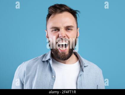 Arrabbiato ragazzo che grida in rabbia, esprimendo la sua aggressione sullo sfondo blu dello studio Foto Stock