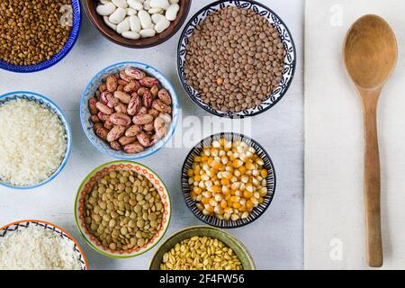 Un closeup di ciotole di cereali assortiti e fagioli su una superficie bianca con un cucchiaio di legno Foto Stock