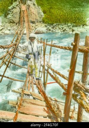 Scatto a tutta lunghezza di J H Arthur, attraversando un ponte di corda dilapidata in una giornata di sole, con scatole di attrezzature appese intorno al suo collo, provincia di Sichuan, Cina, 1917. Dalla collezione di fotografie di Sidney D. Gamble. () Foto Stock