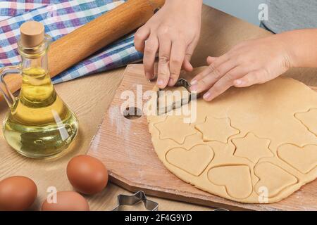 Le mani del bambino tengono una taglierina metallica del biscotto e tagliano i biscotti fatti in casa di varie forme dalla pasta frolla. Su tavola, uova, girasole Foto Stock