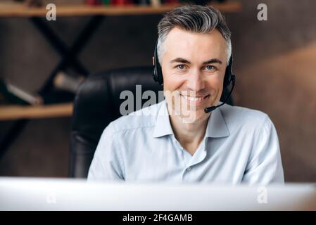 Ritratto di un uomo di mezza età, manager, assistente o addetto al call center, che si siede in un ufficio moderno, indossando una camicia e delle cuffie eleganti, guardando la fotocamera sorridente e amichevole Foto Stock