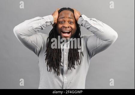 Ritratto di un giovane afro-americano scioccato con dreadlock che indossa una camicia guardando la macchina fotografica a sorpresa, stordito con una storia incredibile, tenendo le mani sulla testa, emozioni di espressioni umane Foto Stock