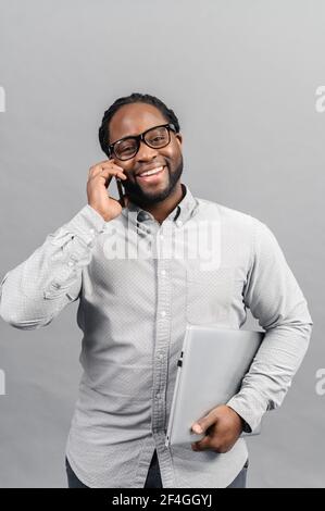 Concetto di ricevere le buone notizie. Felice afro americano uomo d'affari in camicia sorridente che parla per telefono con il cliente dopo aver lavorato con il laptop che tiene in mano, si erge su sfondo grigio Foto Stock