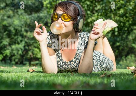 Happy girl con le cuffie ascolta la musica e gonfia una gomma da masticare. Giovane donna elegante in stile casual e occhiali da sole gialli all'aperto Foto Stock