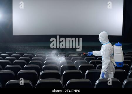 Uomo o donna in indumenti anti-virus protettivi sedili di pulizia in sala cinema con disinfettanti. Concetto di preparazione sala cinema per nuovi visitatori. Foto Stock