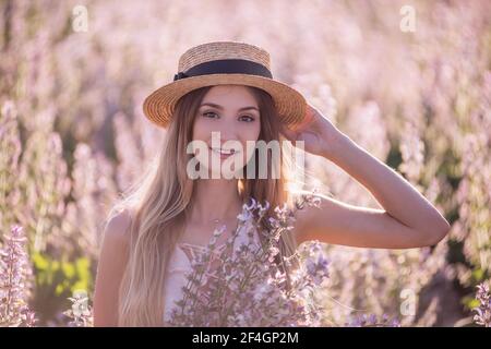 Giovane bionda donna in cappello di paglia sullo sfondo del campo fiorente di salvia rosa. Primo piano ritratto bella ragazza che tiene fiori bouquet in mani. W Foto Stock