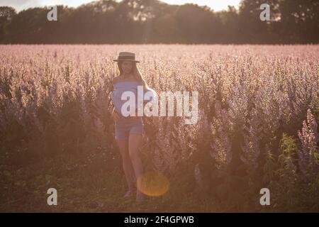 Una giovane donna con camicia blu e pantaloncini in denim si erge su un campo di salvia rosa in fiore. Ritratto di ragazza con lunghi capelli biondi che indossa un cappello di paglia al sole Foto Stock