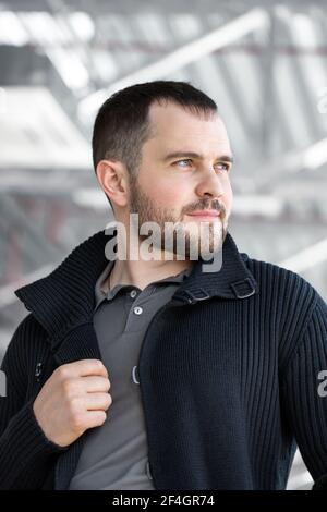 Portret un bel giovane uomo con un elegante taglio di capelli e barba che guarda fuori dalla finestra. Immagine di un uomo bruna felice che indossa abiti formali sopra bac grigio Foto Stock