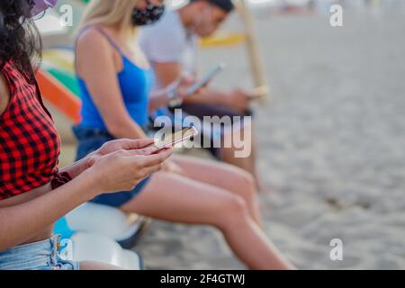 Gli amici seduti in spiaggia e che usano i loro smartphone in social distanza e con maschera facciale attivata Foto Stock