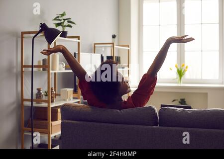 Vista posteriore della giovane donna felice che allunga le braccia seduta sul divano in soggiorno Foto Stock