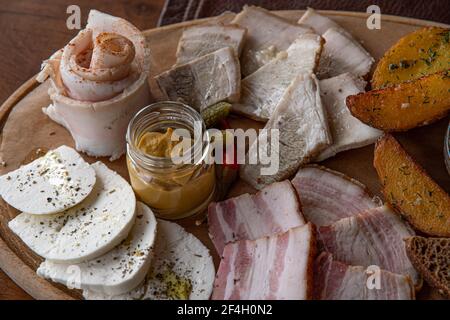 Vassoio di legno per closeup con carne assortita, grasso di maiale, fette di pancetta e antipasti rustici di formaggio con patate arrostite e senape in un piccolo vasetto di vetro. Locale Foto Stock