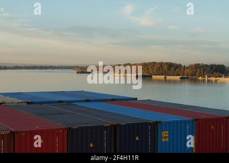 Chiatte fluviali e barche in rimorso ormeggiate vicino al terminal dei container sul fiume Columbia circondato da alberi e cespugli sotto il cielo blu. Foto Stock